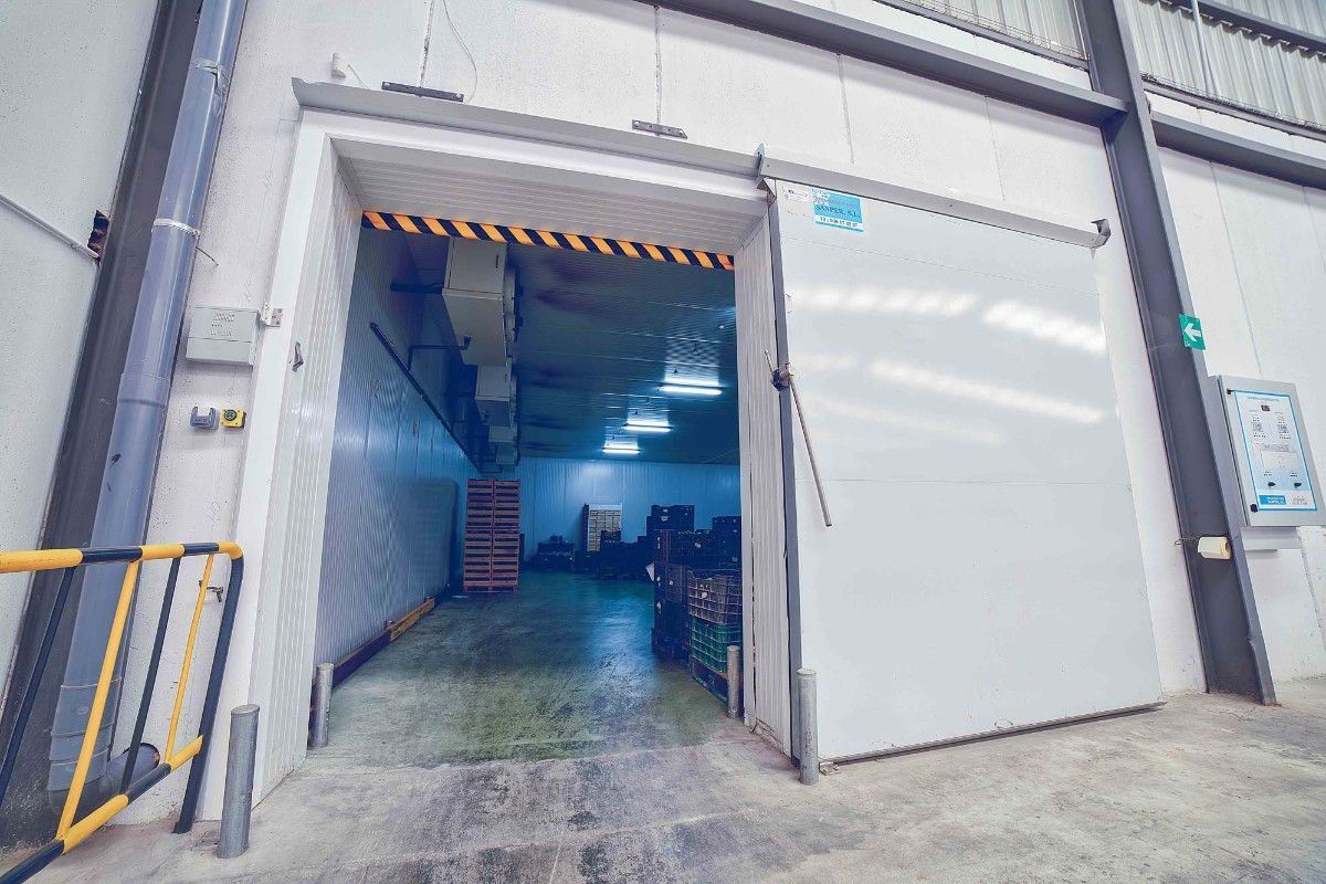 La entrada de un almacén con una puerta corredera blanca abierta deja ver una zona de almacenamiento bien iluminada con mercancías apiladas en su interior.