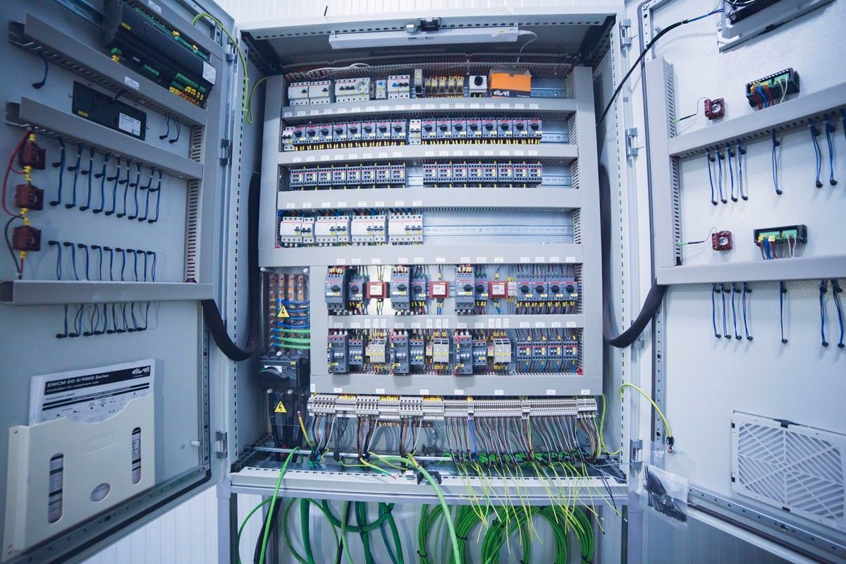 Un panel de control eléctrico industrial abierto con varios componentes, cableado y bloques de terminales montados sobre rieles grises.