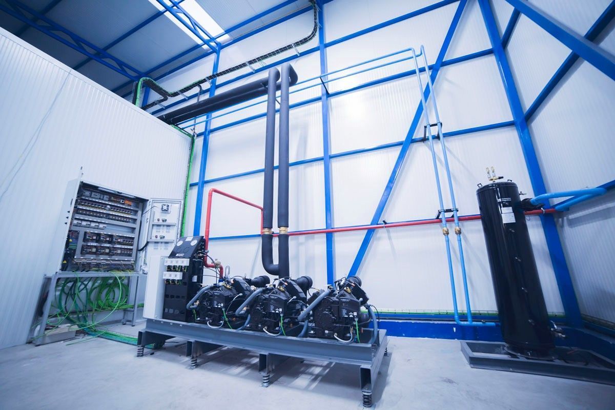 Un bastidor para compresores de refrigeración en un edificio industrial con pared blanca, estructura metálica azul y panel eléctrico.