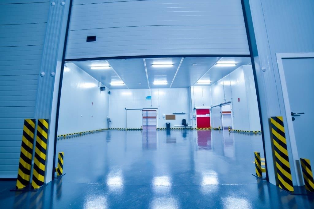 Una cámara frigorífica industrial vacía y muy iluminada, vista a través de una puerta de almacén abierta con postes de seguridad amarillos y negros.