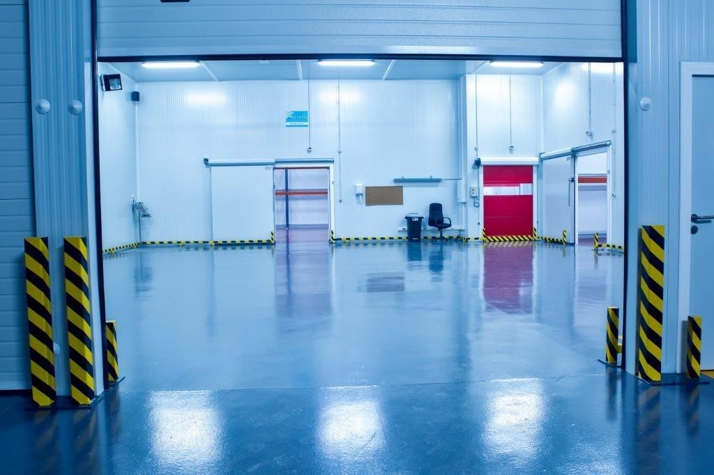 Vista de un espacio industrial aséptico, como un almacén, con paredes blancas, suelo azul pulido y postes de seguridad amarillos y negros.