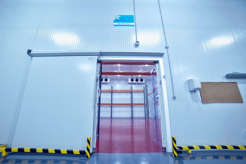 Se abre la puerta a una cámara frigorífica comercial con estanterías y suelo rojo.