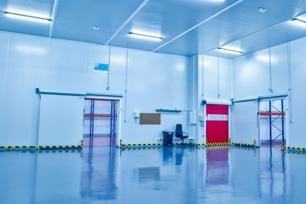 Una cámara frigorífica industrial muy iluminada, con suelos azules, paredes blancas, estanterías y una puerta roja cerrada.