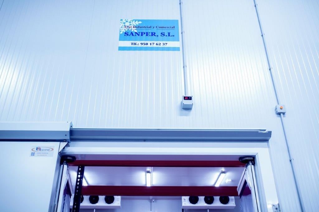 Una gran puerta frigorífica industrial con una pared de paneles metálicos blancos acanalados y un letrero azul de la empresa sobre la entrada.