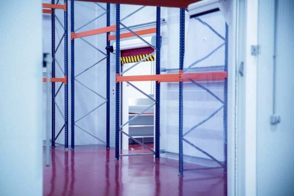 El interior vacío de un almacén industrial con estanterías metálicas azules, vigas de soporte naranjas y un suelo reflectante rojo.