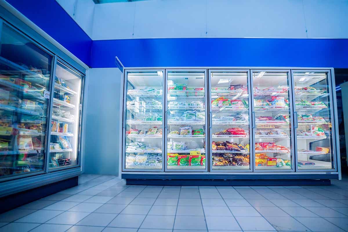 Grandes congeladores de cristal repletos de alimentos en un luminoso pasillo de supermercado alicatado, con una destacada sección de pared azul.