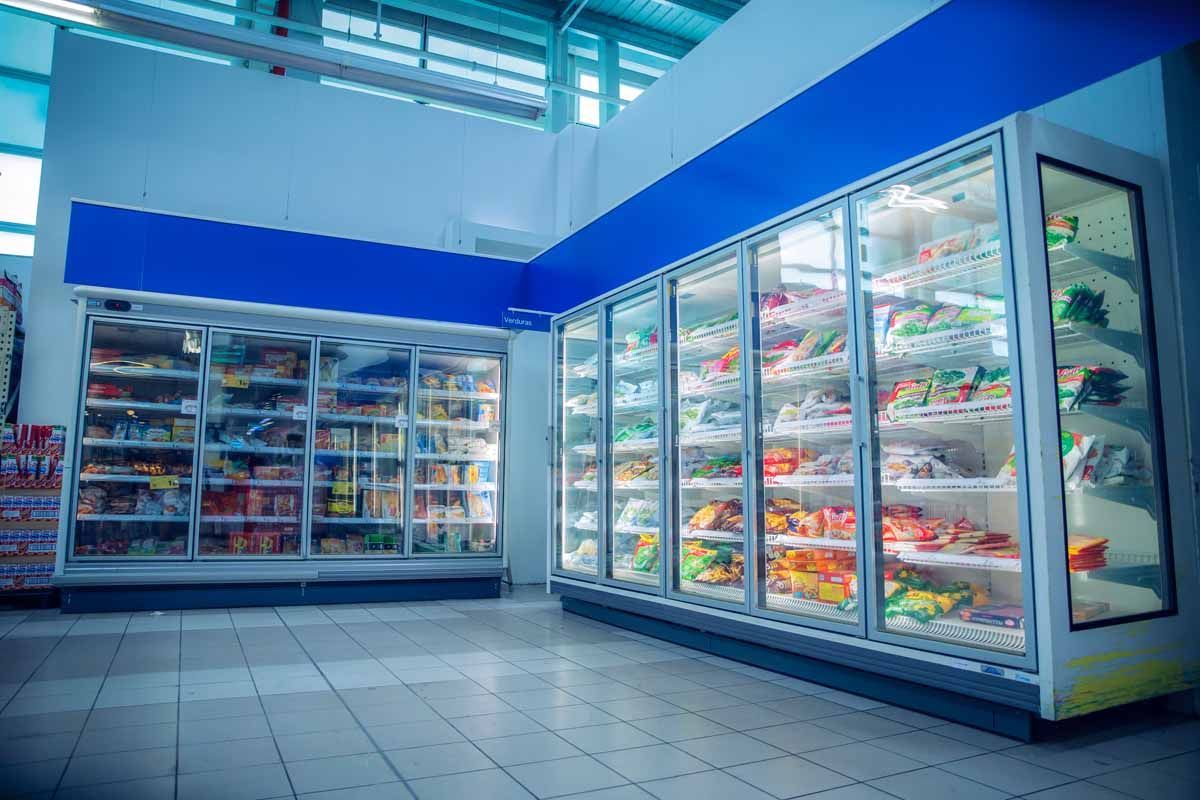 Filas de refrigeradores con puertas de vidrio, abiertos hasta el borde, que contienen productos de alimentación en un supermercado bien iluminado con suelos de baldosas.