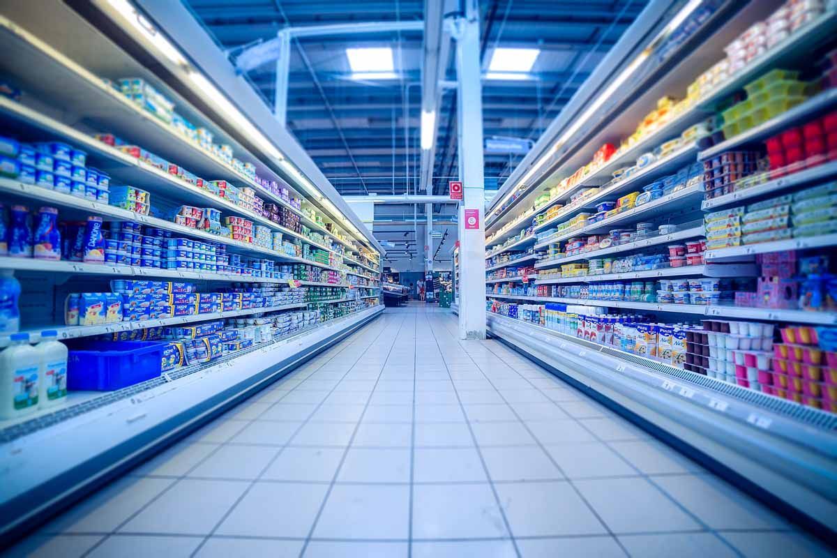 Vista simétrica de un pasillo de supermercado con largas estanterías refrigeradas repletas de diversos productos de alimentación.