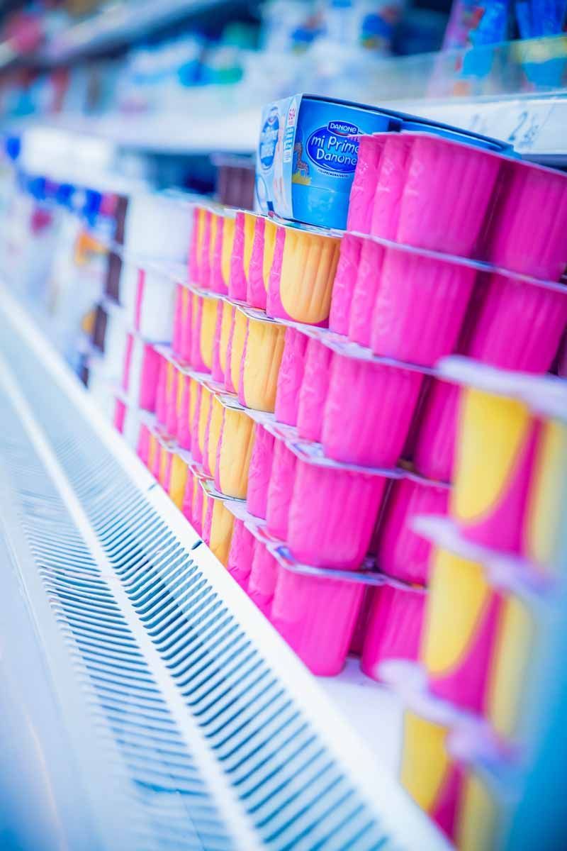 Filas de vasitos de yogur rosas y amarillos expuestos en una estantería refrigerada de un supermercado.