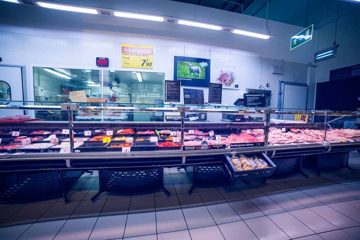 Un mostrador de carne en un supermercado con exhibiciones de cortes crudos bajo una brillante iluminación cenital.
