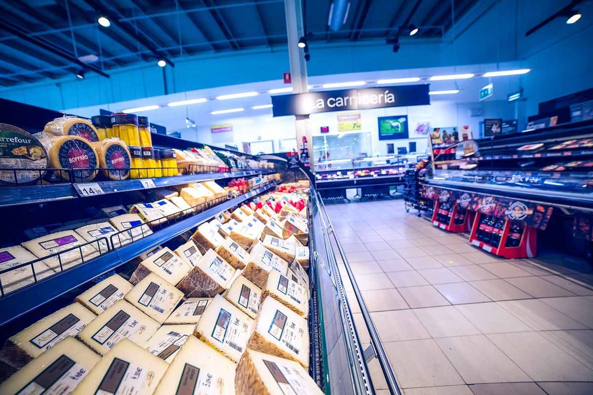 Pasillo de supermercado con estantes repletos de quesos envasados ​​de diversos tipos, que conduce hacia un mostrador de carnes al fondo.