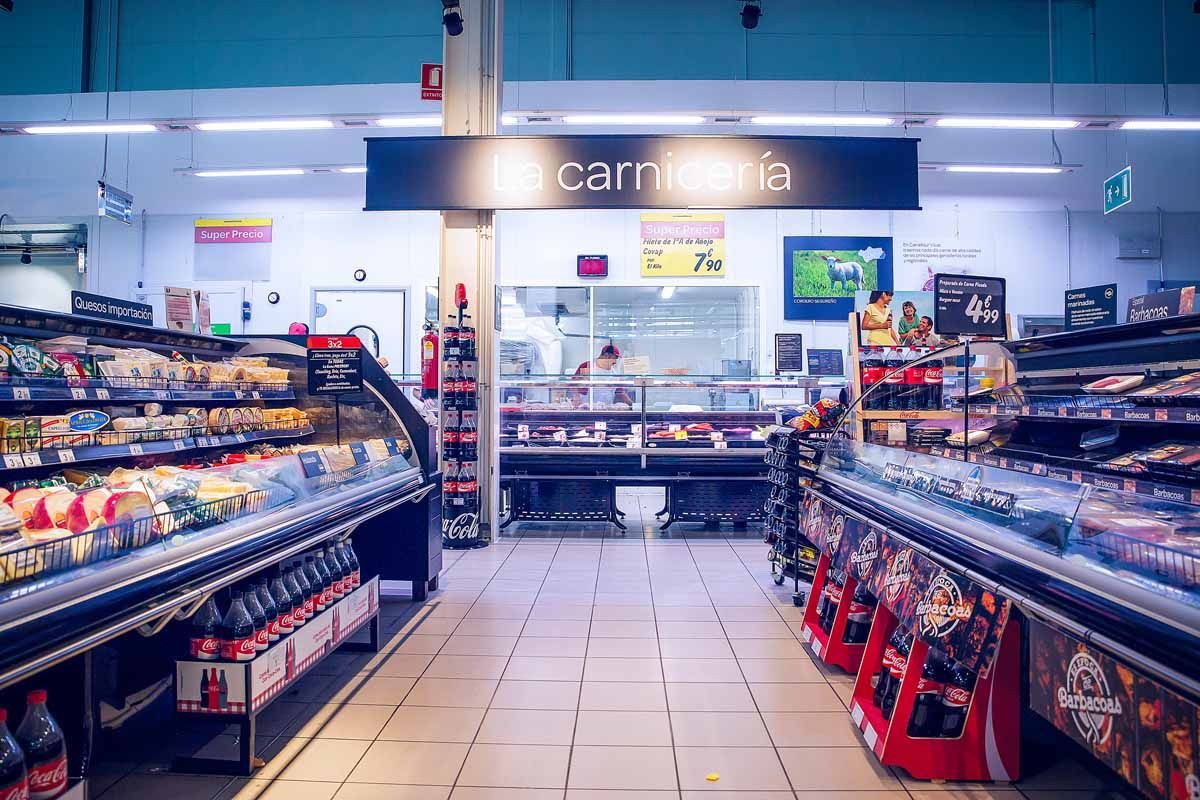 Vista de un pasillo de supermercado hacia un mostrador de charcutería con la etiqueta 