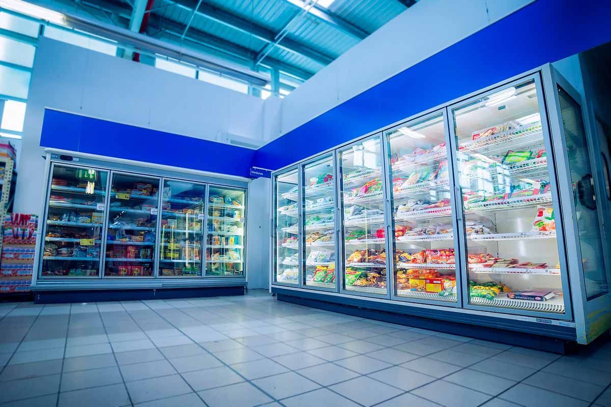 Grandes y modernas cámaras frigoríficas de supermercado con puertas de cristal, repletas de diversos productos alimenticios.