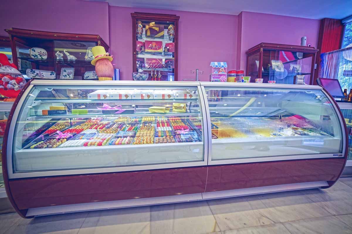 Una vitrina de cristal curvada llena de coloridos dulces y golosinas dentro de una tienda con paredes rosas y estanterías de madera oscura.