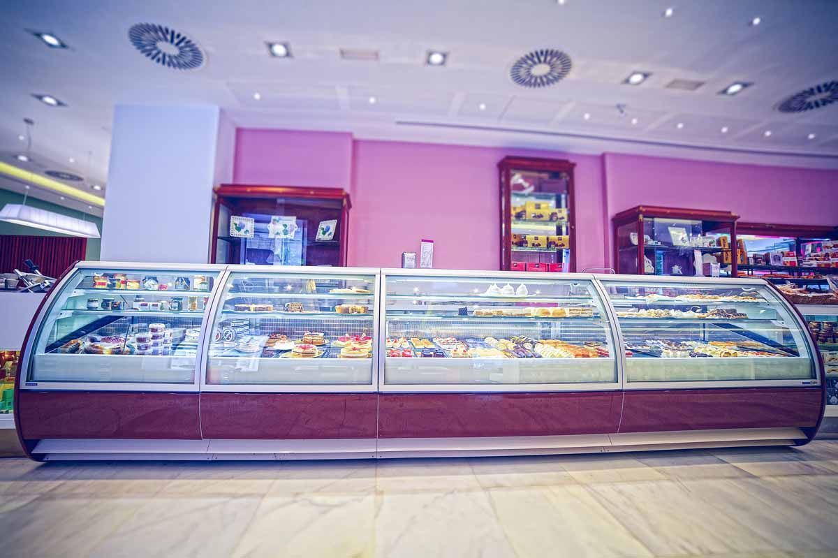 Una vitrina larga y curvada de cristal, repleta de pasteles y tartas, en una panadería con paredes rosas e iluminación brillante en el techo.