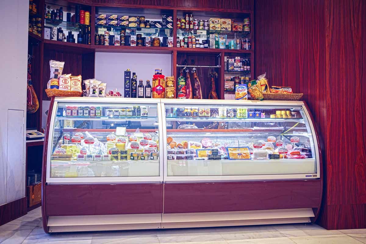 Una vitrina refrigerada de charcutería llena de alimentos, con estantes rematados con productos, en una tienda con paneles de madera.