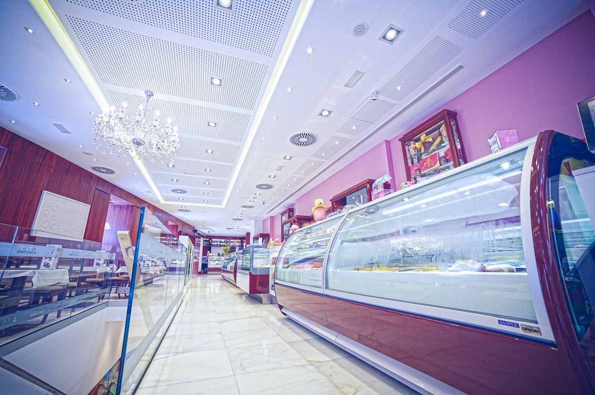 Una heladería luminosa y moderna con paredes rosas, un largo mostrador de cristal, suelos de baldosas y luces decorativas en el techo.