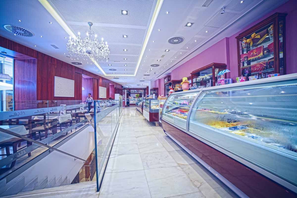 Una elegante pastelería con una larga vitrina de cristal, suelo de baldosas blancas, modernas lámparas de araña y paredes rojas.