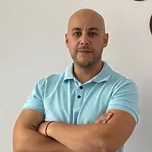Un hombre calvo con barba está de pie con los brazos cruzados frente a una pared blanca.