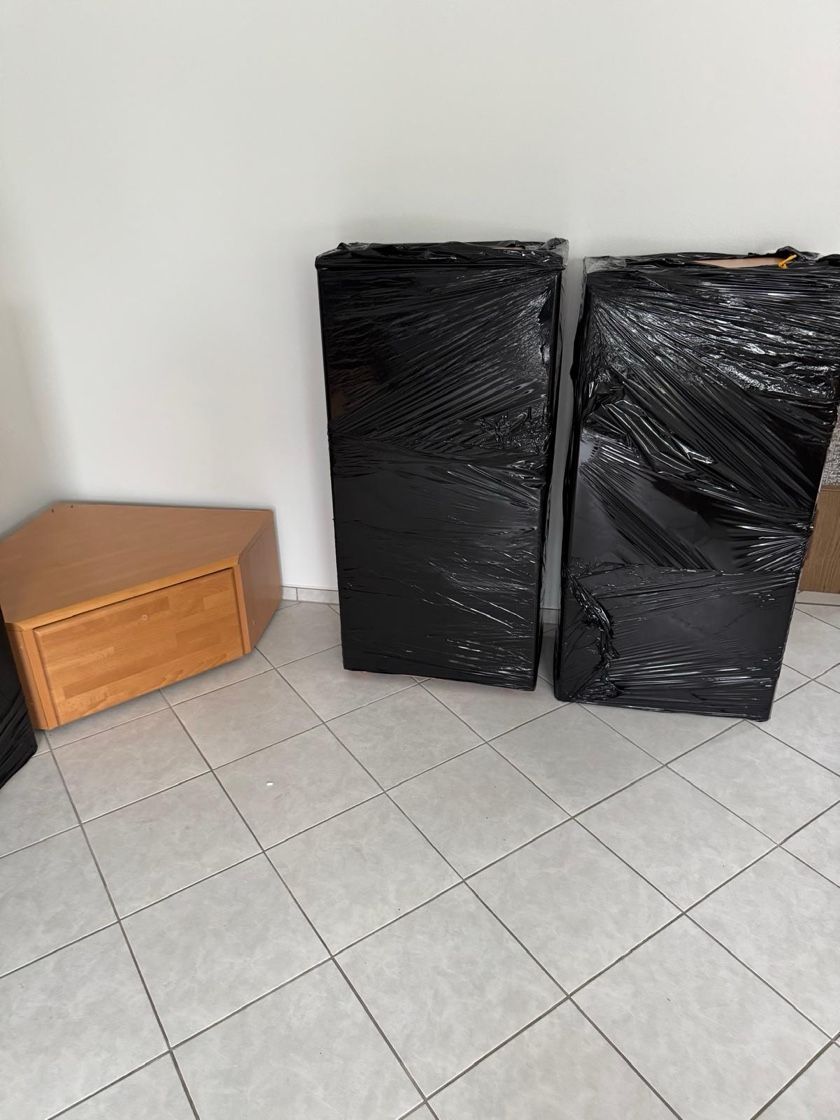 Zwei schwarz verpackte rechteckige Objekte und eine Holzkommode stehen auf einem gefliesten Boden neben einer weißen Wand.