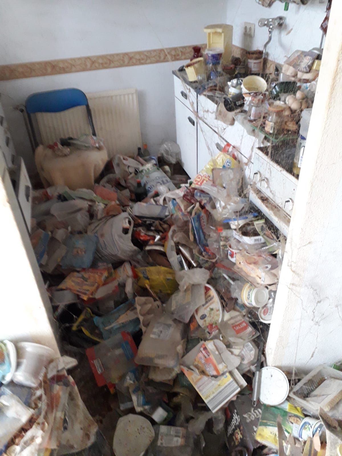 Eine überfüllte Küche mit Müllhaufen auf dem Boden und den Arbeitsflächen, einem Stuhl und einem Schrank ist zu sehen.