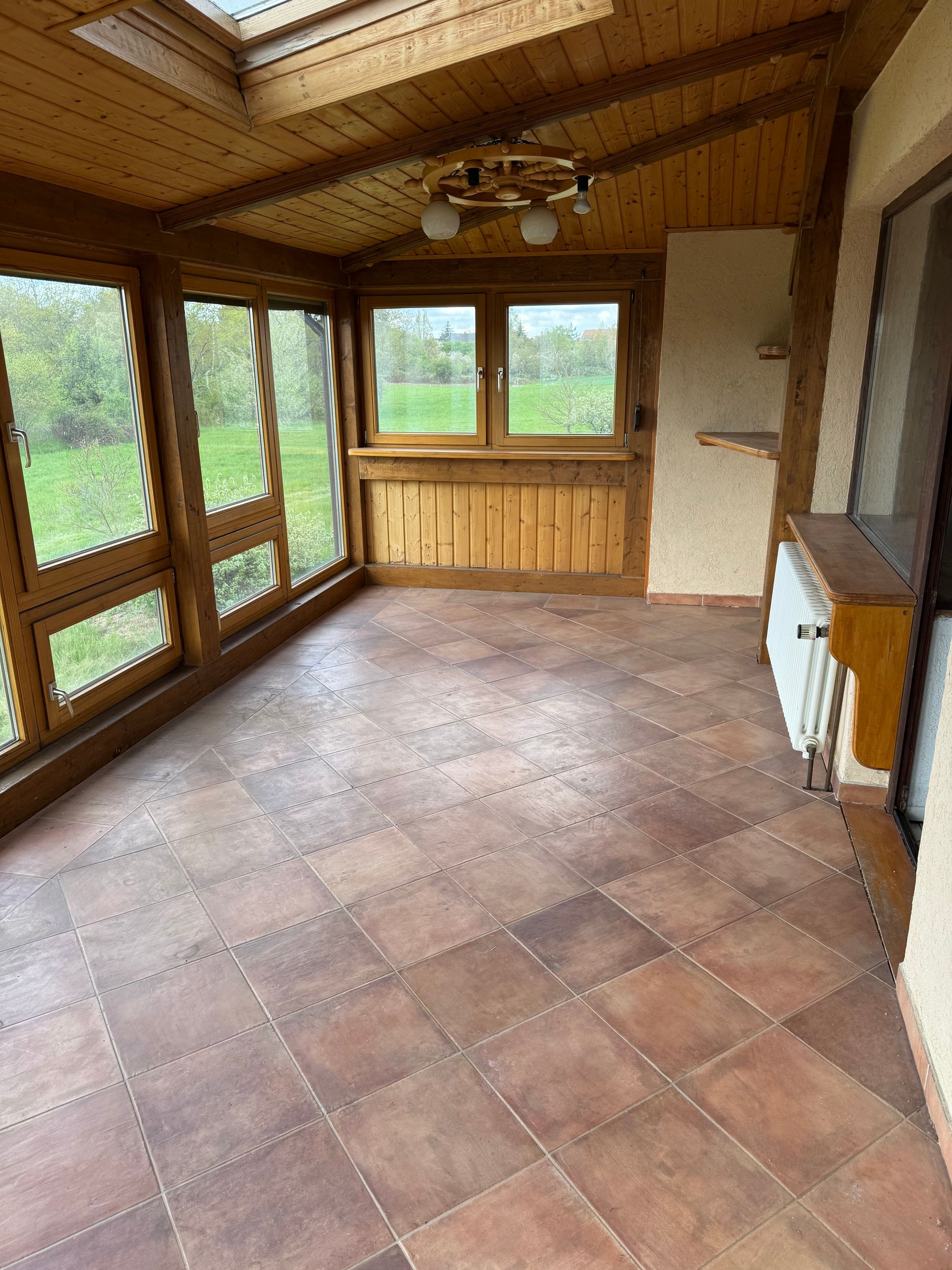 Wintergarten mit Holzvertäfelung, Fliesenboden und Oberlichtern mit Blick auf eine grüne Wiese.