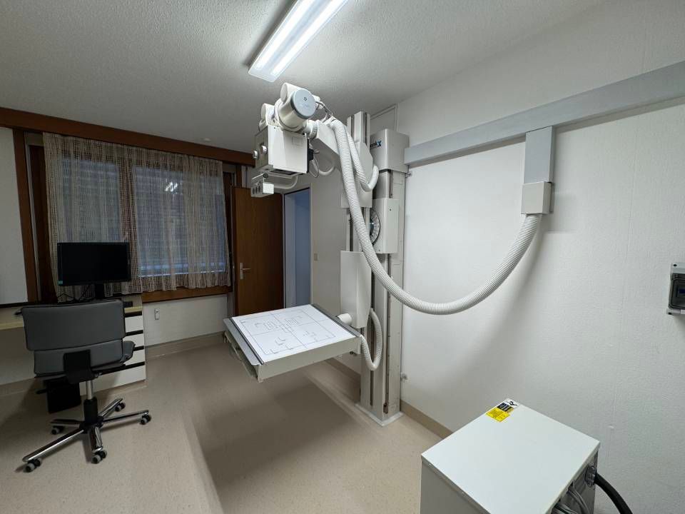Ein Raum mit einem Röntgengerät mit hängendem Detektor, einem Schreibtisch mit Monitor und einem weissen Aufbewahrungsschrank.