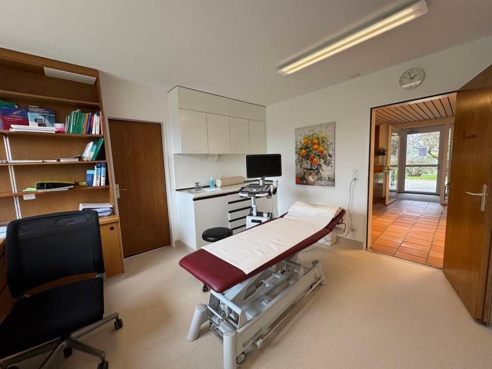 Ein Untersuchungsraum mit verstellbarer, bordeauxroter Untersuchungsliege, Monitor, Schreibtisch, Regalen und offener Tür.