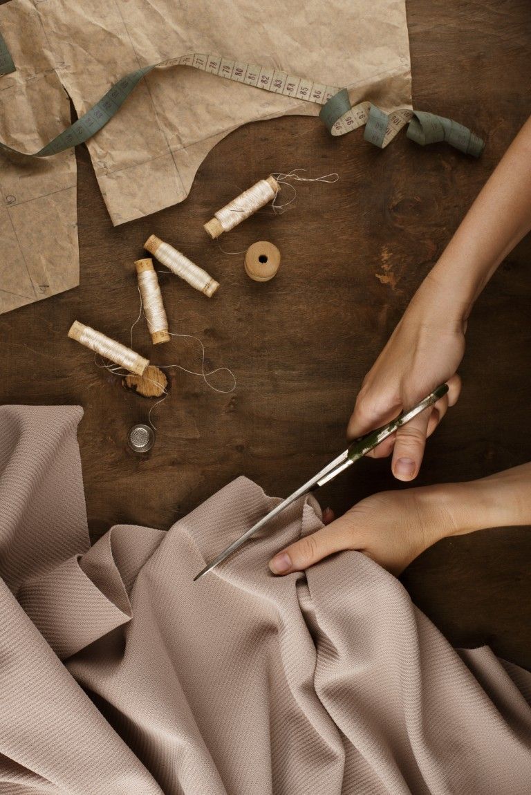 Manos cortando tela beige sobre una mesa de madera con herramientas de costura y trozos de tiza.