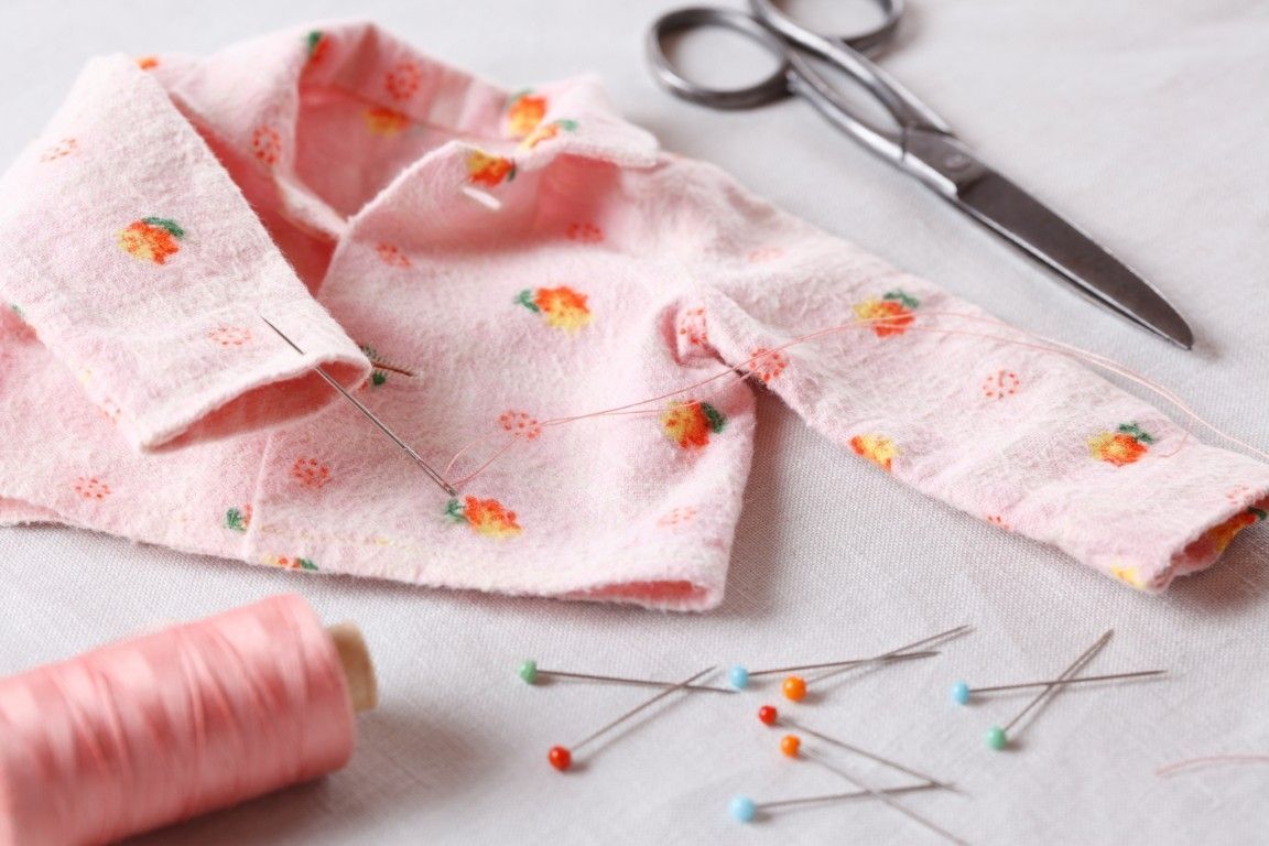 Tela rosa con estampado floral, tijeras, carrete de hilo y alfileres dispuestos para coser sobre una superficie blanca.