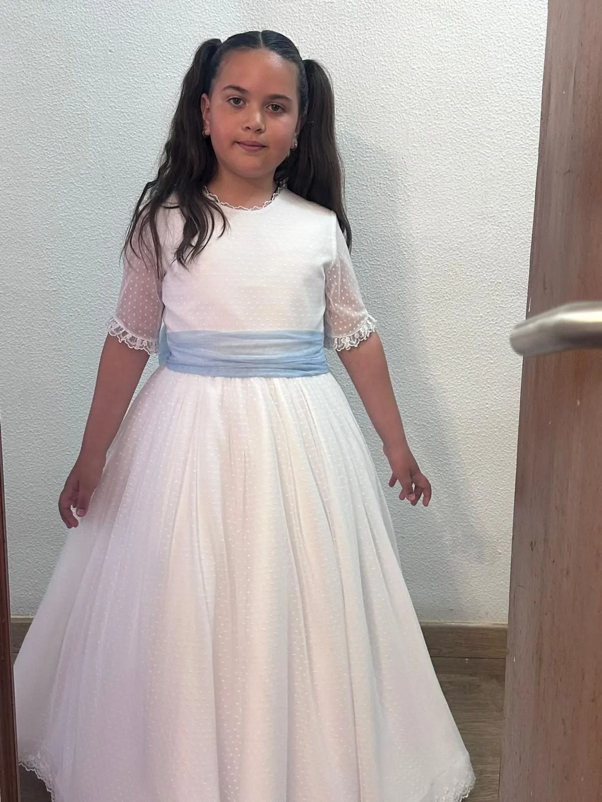 Niña con un vestido blanco de gala y fajín azul, de pie en el interior de una casa, apoyada contra una pared texturizada.