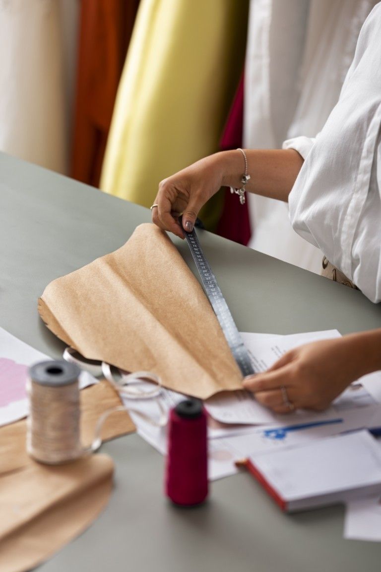 Una persona mide tela beige con una regla en una mesa de trabajo, con carretes de costura y papeles cerca.