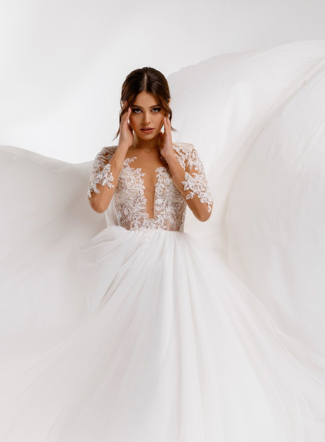 Unas manos sujetan la parte trasera de un vestido de novia de encaje blanco junto a una ventana luminosa.
