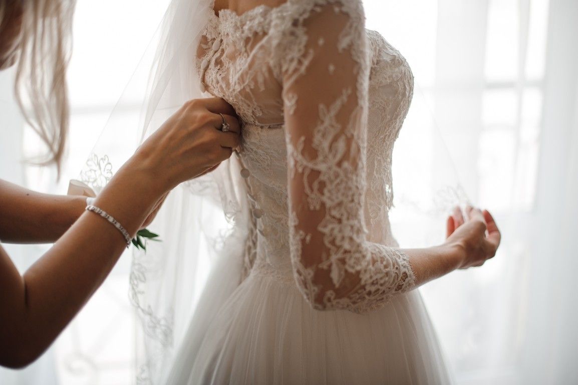 Unas manos sujetan la parte trasera de un vestido de novia de encaje blanco junto a una ventana luminosa.