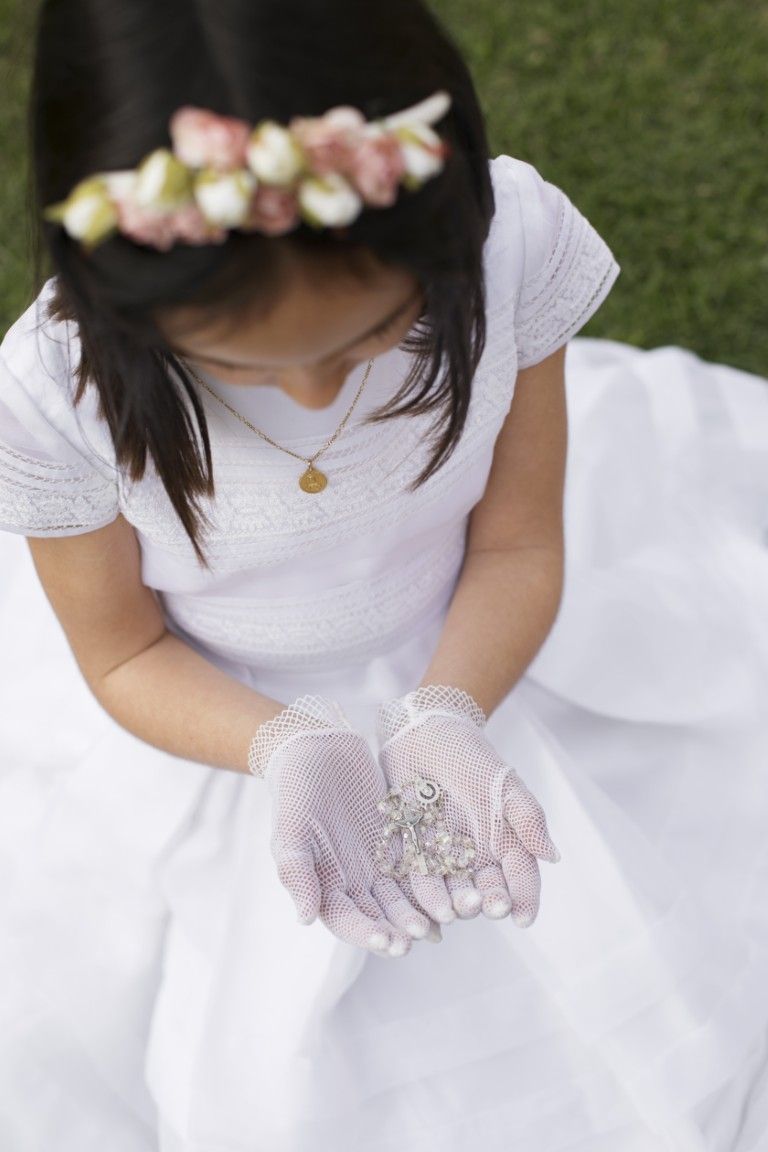 Niña con vestido blanco, corona de flores y guantes de encaje, mirando hacia abajo con las manos juntas al aire libre.