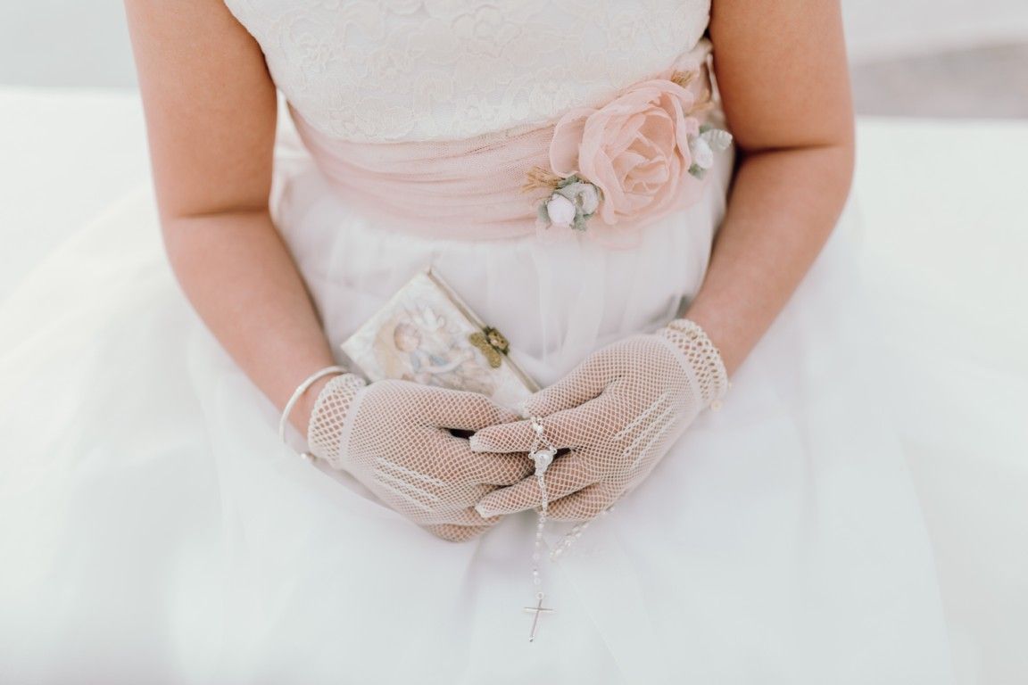 Niña con vestido blanco y fajín rosa sosteniendo un pequeño ramo de flores y guantes de encaje.