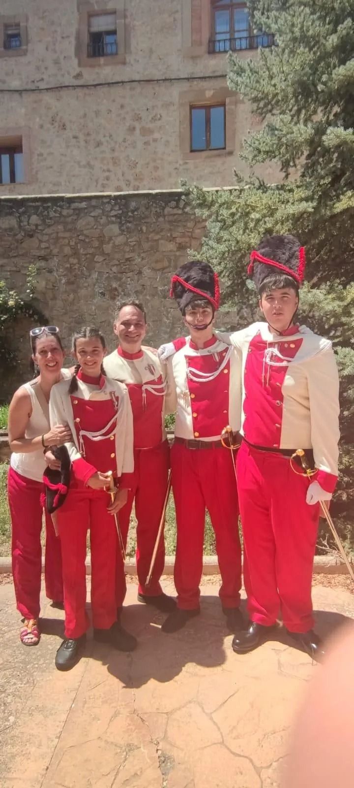 Cinco personas vestidas con trajes tradicionales rojos y blancos posan al aire libre junto a un edificio de piedra.