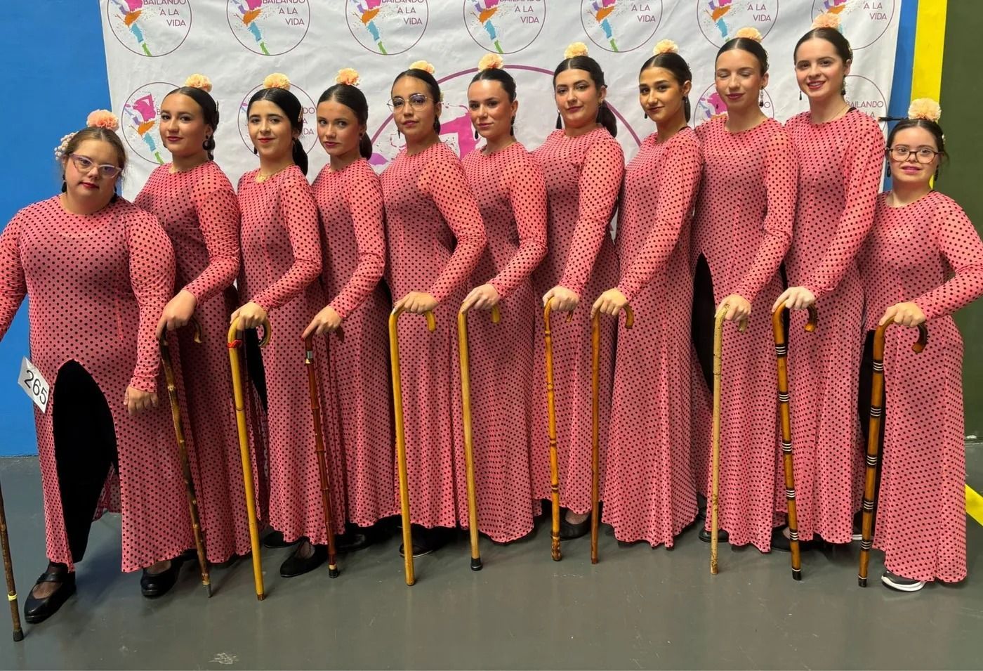 Un grupo de artistas con trajes rosas estampados a juego, sosteniendo bastones, se colocan en fila sobre el escenario.
