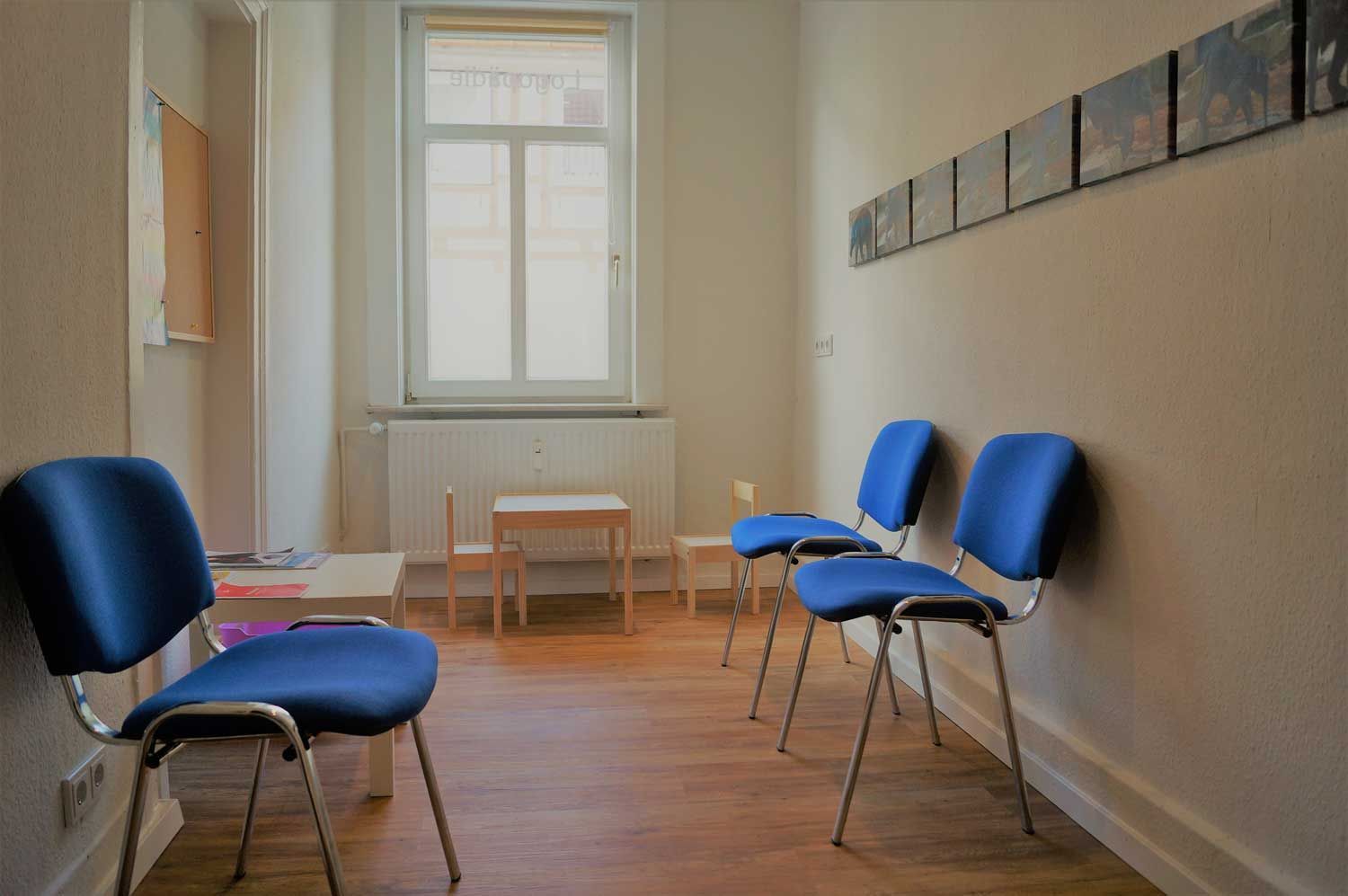 Wartezimmer mit blauen Stühlen, kleinen Tischen und einem Fenster.