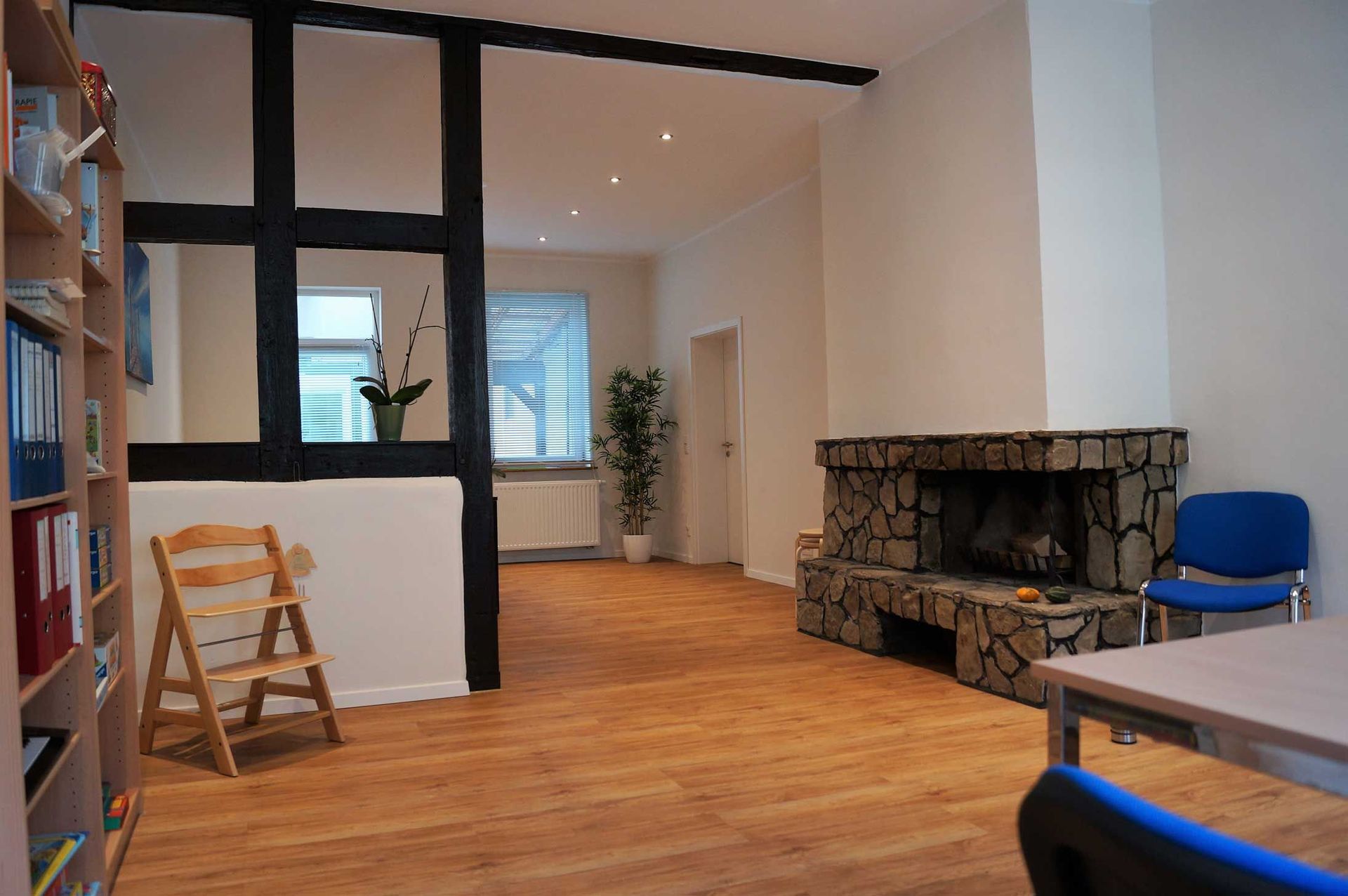 Zimmer mit Holzboden, Kamin, Bücherregal und dekorativen schwarzen Balken. Ein Tritthocker steht neben den Balken.