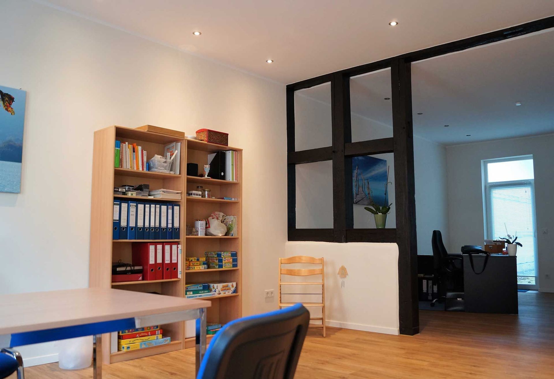 Helles Bürointerieur: Bücherregal, Schreibtisch und eine dunkel gerahmte, offene Tür, die in einen anderen Raum führt.