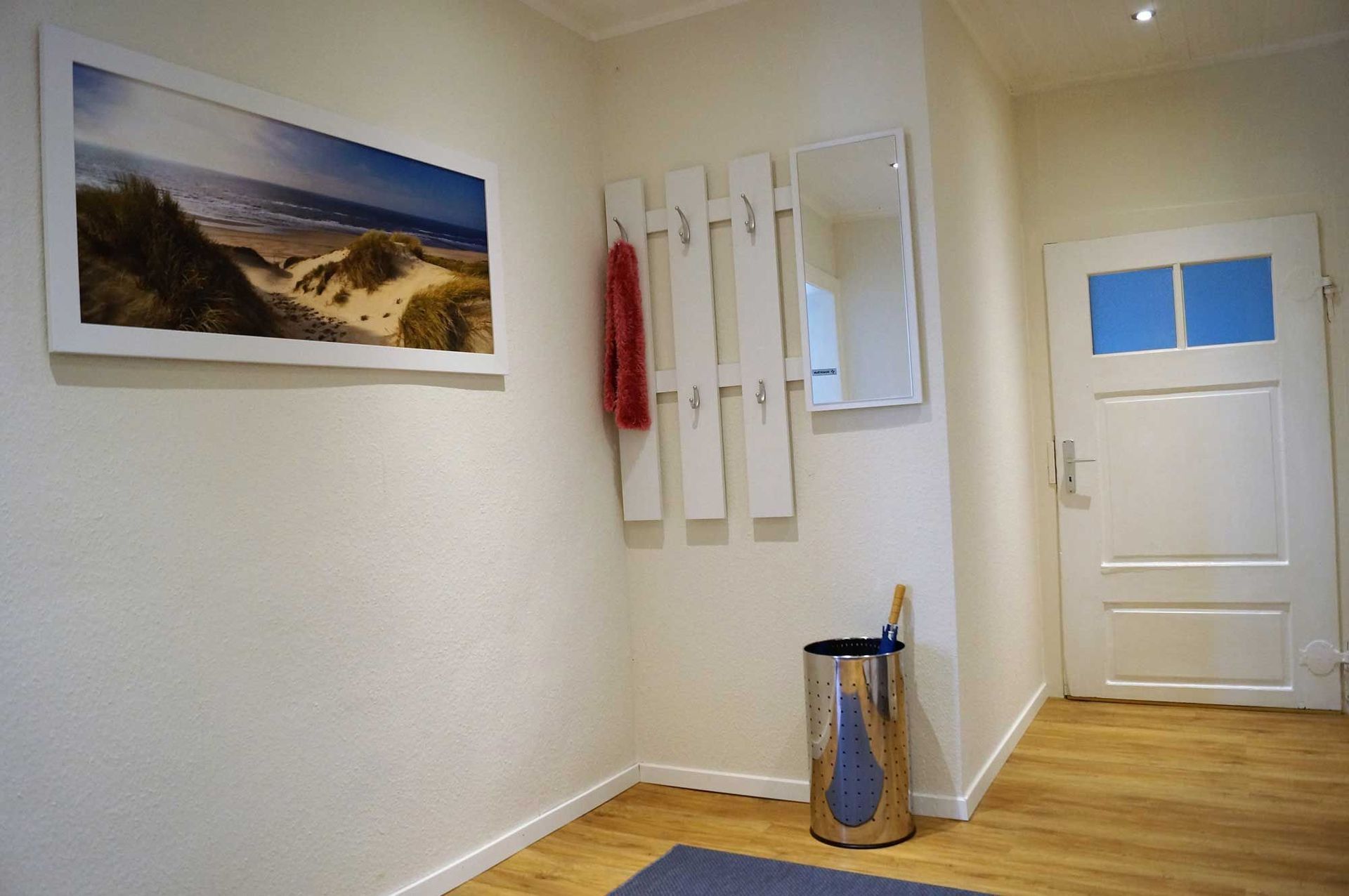 Flur mit Strandfoto, weißer Garderobe, Spiegel und weißer Tür mit blauem Fenster.