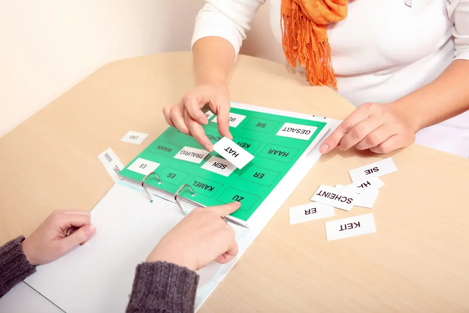 Zwei Personen sitzen an einem Tisch und interagieren mit einem grünen Spielbrett und Wortkarten. Hände zeigen auf Karten und platzieren diese.