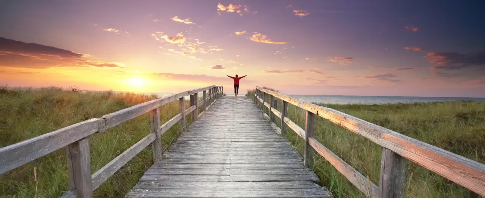 Eine Person mit ausgestreckten Armen steht auf einem Holzsteg und blickt auf einen leuchtenden Sonnenuntergang über Wasser und grasbewachsenen Dünen.