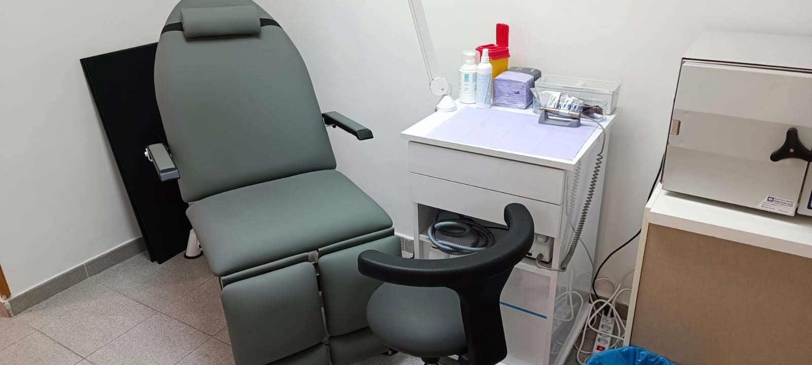 Pequeño consultorio médico con una silla de exploración color verde azulado, un taburete con ruedas y suministros sobre una mesa auxiliar.
