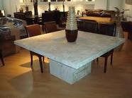 Mesa de comedor cuadrada de mármol con un gran jarrón, rodeada de sillas en una sala de exposición.