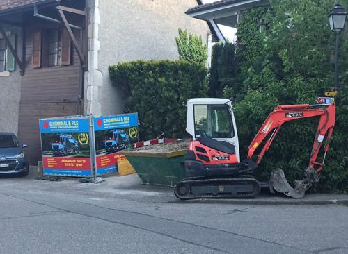 Une petite excavatrice est garée sur le bord de la route à côté d'une benne à ordures.