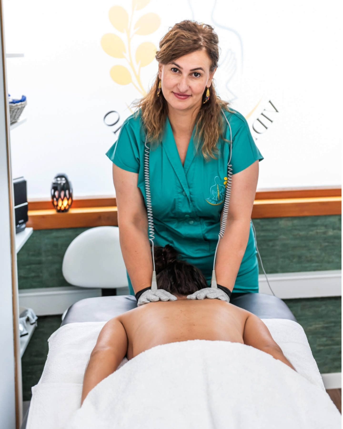 Mujer con uniforme verde aplica terapia eléctrica a un cliente acostado boca abajo en una mesa de masajes.
