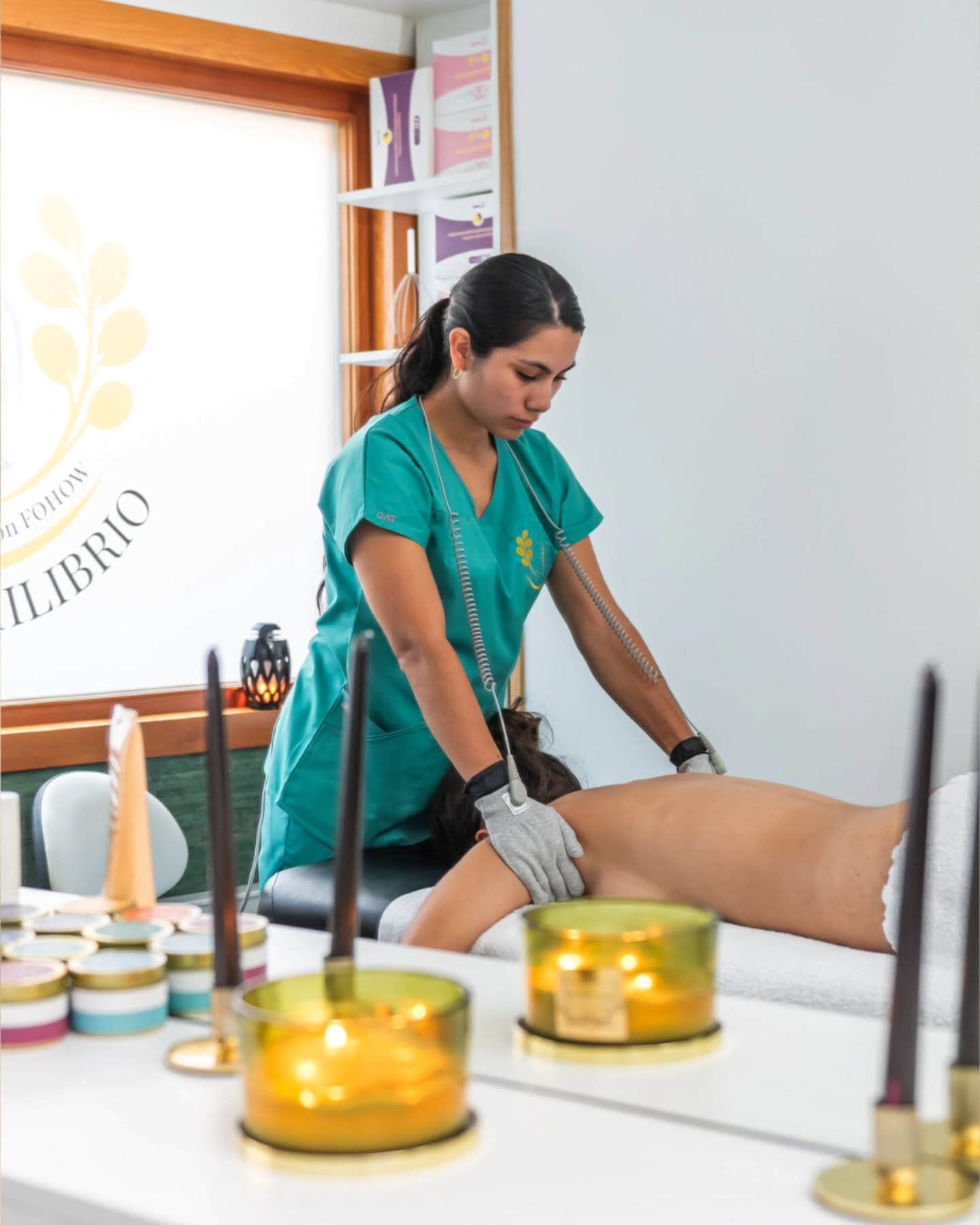 Una mujer con uniforme verde azulado realiza terapia a una persona tumbada en una camilla de masaje. Velas en primer plano.