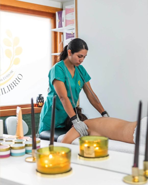 Una mujer con uniforme verde azulado realiza terapia a una persona tumbada en una camilla de masaje. Velas en primer plano.